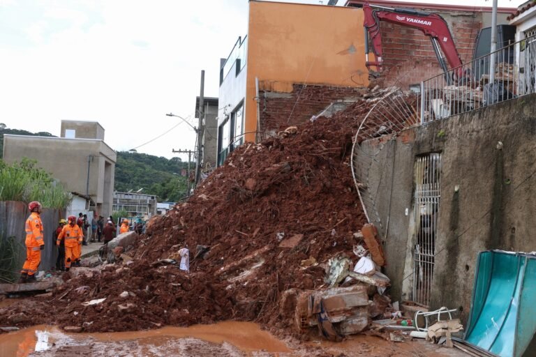credito extraordinario: Deslizamento de terra em área urbana com agentes de resgate e escavadeira atuando entre escombros.