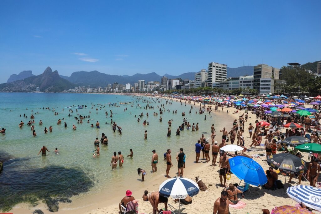 Multidão de pessoas em uma praia ensolarada com guarda-sóis coloridos, prédios ao fundo e montanhas sob um céu azul.
