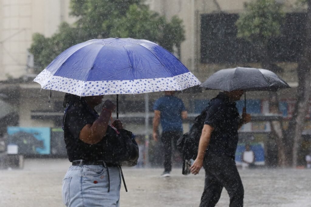 previsão do tempo: pessoas caminham sob chuva forte com guarda-chuvas em área urbana