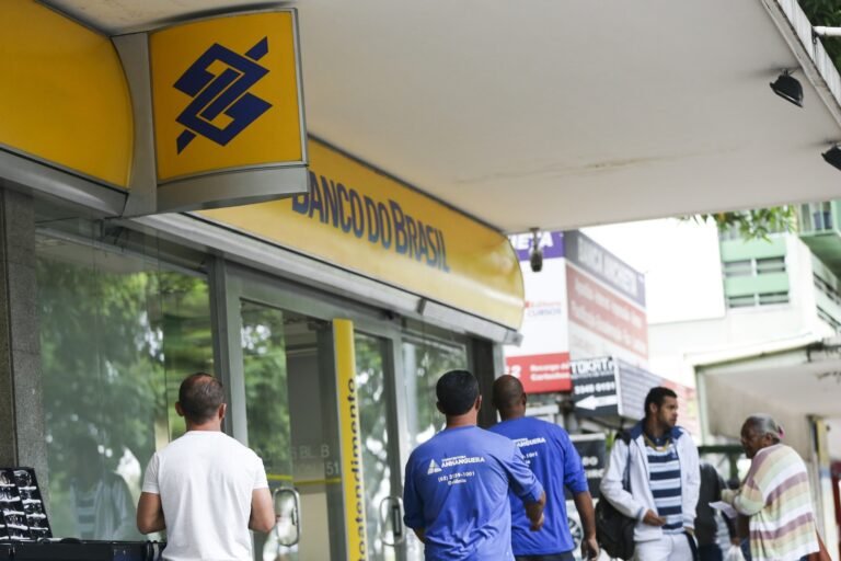 Pessoas caminham na calçada em frente a uma agência do "Banco do Brasil".