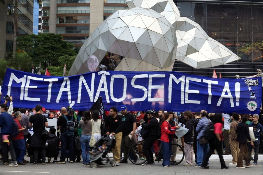 Manifestantes seguram faixa com os dizeres "META, NÃO SE META!" em protesto na rua.